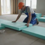 Ouvrier posant des panneaux isolants XPS sur une dalle béton lors de travaux d'isolation thermique des sols, lumière naturelle de chantier résidentiel